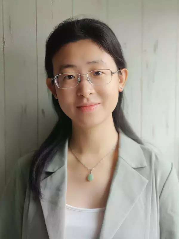 Professor Xia smiles gently at the camera. She is wearing a pale green-gray blazer with a white shirt underneath. Her medium length hair is half up and half down. She is wearing a necklace with a silver chain and pale green stone. Behind her is wood painted pale green.